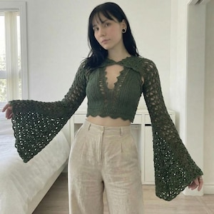 May include: A dark green crochet top with long, flared sleeves. The top has a scalloped neckline and a keyhole detail at the chest. The sleeves are open-weave, and the top is paired with beige pants.