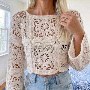 May include: A cream-coloured crochet top with a square neckline and bell sleeves. The top features a floral pattern and is paired with blue denim jeans. The crochet design is open, creating a textured appearance.