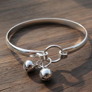 Peut inclure: Un bracelet jonc argenté avec deux petits grelots ronds. Le bracelet a un design simple et élégant avec un fermoir circulaire. Les cloches sont suspendues à une petite chaîne, ajoutant une touche délicate. Le bracelet est présenté sur une surface en bois.