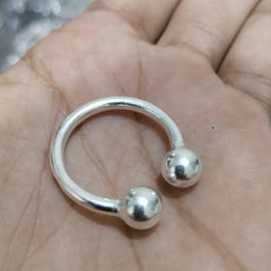 Puede incluir: Un anillo de color plateado en forma de herradura con un acabado liso y pulido, que presenta dos extremos esféricos. La banda abierta tiene una forma circular y posee una superficie metálica reflectante que capta la luz con brillo.