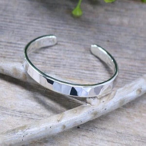 Op de afbeelding: Een zilveren manchetarmband met een facetontwerp. De open armband is gemaakt van glanzend metaal en rust op een lichtgekleurd houten oppervlak. Het geometrische patroon van de armband reflecteert licht.