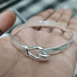 Peut inclure: Un bracelet jonc en argent poli doté d'un fermoir de style crochet. L'anneau circulaire est lisse et réfléchissant, avec un détail décoratif en fil enroulé positionné juste avant la fermeture à crochet entrelacée.