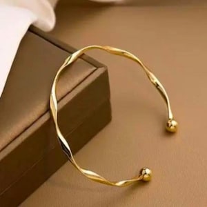May include: A gold-toned open cuff bracelet featuring a twisted, wavy metal band. The bracelet ends in two small, smooth, spherical ball terminals at each opening, creating a continuous, reflective, and polished metallic surface.