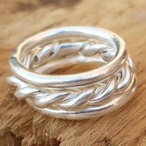 May include: A silver ring composed of three interlocking bands. Two of the bands feature a smooth, polished finish, while the central band displays a twisted, rope-like texture. The metal has a bright, reflective surface throughout the design.