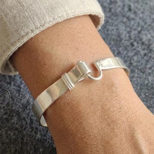 Może przedstawiać: Srebrna bransoletka typu mankiet z zapięciem na haczyk. Bransoletka wykonana jest z polerowanego metalu i ma prosty, elegancki design. Zapięcie na haczyk to unikalny detal. Bransoletka jest noszona na nadgarstku.