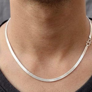 Puede incluir: Un collar de cadena tipo espiga en tono plateado con un diseño de eslabones planos y flexibles que descansa sobre el cuello. El collar incluye un mecanismo de cierre en forma de S visible en el lado derecho, uniendo los segmentos metálicos.