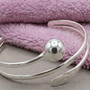 May include: A silver-toned open cuff bracelet featuring a polished spherical ball at one end and a flat, tapered end with a subtle engraved pattern. The bracelet has a bypass design that wraps around the wrist with a smooth, reflective metallic finish.