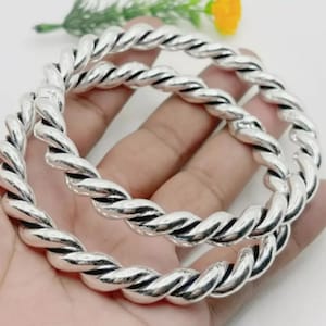 May include: Two silver-coloured bangles in a twisted rope design. The bracelets are made of a shiny metal and have a textured, braided appearance. The bangles are shown against a neutral background.