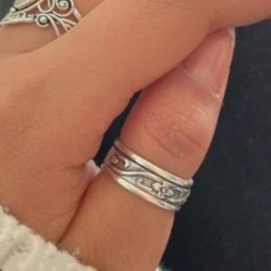 以下が含まれることがあります： 中央に酸化仕上げの施された花やスクロール模様のエンボス加工が繰り返されているシルバーのバンドリング。リングの上下には滑らかで光沢のある縁取りがあり、テクスチャーのある中央部分を囲むようにデザインされています。