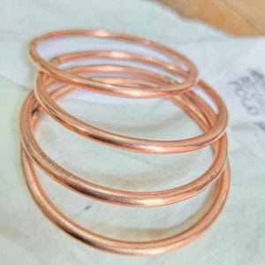 May include: A set of four stacked circular bangles made of polished copper. Each bangle features a smooth, rounded surface and a consistent metallic finish, creating a simple, uniform appearance when layered together in a vertical arrangement.
