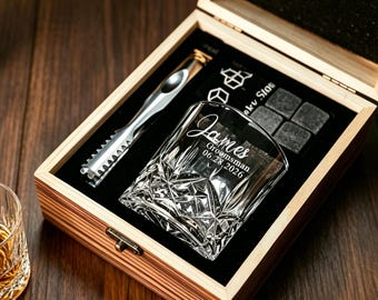 May include: A wooden gift box containing a clear crystal whiskey glass with the etched text James, Groomsman, 06.28.2026, a silver metal ice tong, and four square dark grey stone whiskey cubes set into a black foam insert.