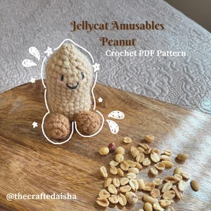 May include: A crocheted peanut toy with a smiling face and two brown peanut halves at the base. The text "Jellycat Amusables Peanut Crochet PDF Pattern" is above the toy. Peanuts are scattered on a wooden board.