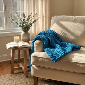 May include: A chunky knit throw blanket in a vibrant teal blue color draped over a beige upholstered armchair. The blanket features a thick, soft yarn texture with large, visible loops created by a hand-knitted technique, providing a cozy appearance.