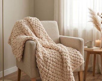 May include: A chunky knit blanket in a cream color draped over a beige armchair. The blanket has a thick, woven texture. A floor lamp, small side table, and a round rug are also visible in the room. The overall theme is cozy and comfortable.