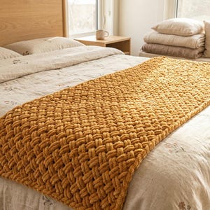 Könnte beinhalten: Eine senffarbene, grob gestrickte Decke liegt auf einem Bett. Die Decke hat eine gewebte Textur und liegt auf einer Bettdecke mit floralem Muster. Ein hölzernes Kopfteil und ein Nachttisch sind im Hintergrund zu sehen.