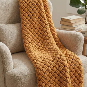 Könnte beinhalten: Eine senfgelbe Wohndecke mit einem groben, handgestrickten Korbgeflechtmuster aus dickem, weichem Garn. Die Decke liegt über einem cremefarbenen Polstersessel mit strukturierter Oberfläche und zeigt ihre dichten, ineinandergreifenden Schlaufen.