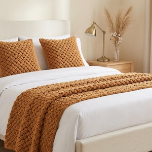 May include: A bed with a cream-colored headboard and frame, featuring white bedding and two square, woven, orange-brown pillows. An orange-brown woven throw blanket is draped across the foot of the bed. A gold lamp and decorative vase are on a wooden nightstand.