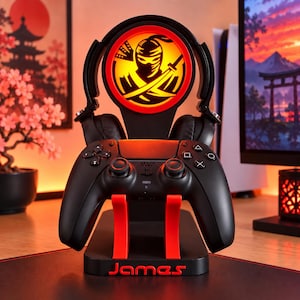 May include: A black gaming controller and headset stand featuring a circular illuminated orange display with a black ninja silhouette graphic. The stand has red support arms and the name James printed in red on the base, holding a black game controller.
