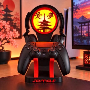 Op de afbeelding: Een zwarte standaard voor een gamecontroller en headset met een cirkelvormig verlicht paneel met een gele gloed, een silhouet van een pagode en Japanse karakters. De zwarte voet heeft rode accenten en de naam James in rode letters op de voorzijde.