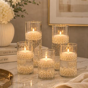 Peut inclure: Cinq vases cylindriques en verre transparent remplis d'eau, de perles artificielles blanches immergées et de bougies piliers blanches flottantes. Les bougies sont allumées, diffusant une lueur chaude à travers le verre et les perles.