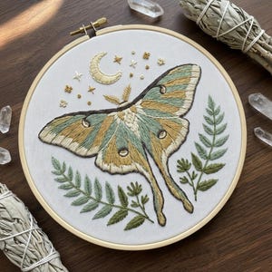 May include: Embroidered artwork featuring a large moth with green and gold wings, surrounded by botanical elements and a crescent moon. The design is set within a round wooden embroidery hoop. The color palette includes shades of green, gold, and cream.