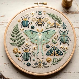 May include: A circular embroidery hoop with a detailed insect and floral design. The central motif is a large, pale green moth with detailed wing patterns. Various beetles, bees, and flowers in green, blue, and yellow surround the moth, all stitched on a neutral fabric.