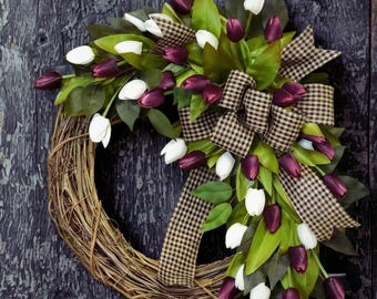 Puede incluir: Una corona con una base de ramas naturales adornada con tulipanes blancos y morados oscuros, hojas verdes y una cinta de tela a cuadros marrones y blancos. El arreglo floral se agrupa en un lado del marco circular de ramas.