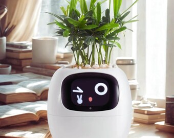 Puede incluir: Una maceta blanca en forma de huevo con una pantalla digital que muestra una cara de dibujos animados y una planta verde. La planta tiene hojas largas y verdes y está plantada con pequeñas piedras blancas. La maceta está sobre una superficie de madera.