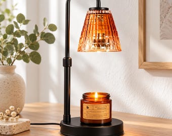 Puede incluir: Una lámpara calentadora de velas de metal negro con brazo ajustable y pantalla de vidrio ámbar texturizado. La lámpara se asienta sobre una base circular, colocada directamente sobre una vela en frasco de vidrio marrón con mecha.
