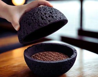 Puede incluir: Un recipiente negro y poroso en forma de cuenco con una tapa que se levanta. El interior del cuenco tiene una superficie texturizada y marrón. El recipiente está sobre una superficie de madera.