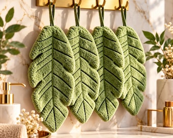 Puede incluir: Cuatro toallas de mano de color verde salvia con forma de hoja hechas de tela texturizada, cada una con una vena central y venas laterales, colgadas de un perchero horizontal de metal dorado con cuatro ganchos en una pared de mármol.