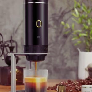 Op de afbeelding: Een zwarte draagbare espressomachine op een zilveren standaard, die koffie in een glas giet. De machine heeft een gouden aan/uit-knop en een klein indicatielampje. Koffiebonen en een kleine gouden beker zijn op de achtergrond zichtbaar.