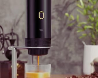 Puede incluir: Una máquina de espresso portátil negra con un soporte plateado, que dispensa café en un vaso transparente. La máquina tiene un botón de encendido dorado y una pequeña luz indicadora. Granos de café y una pequeña taza dorada en el fondo.