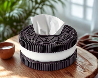 Puede incluir: Una caja de pañuelos redonda diseñada para parecer una galleta tipo sándwich con dos capas exteriores de color marrón oscuro, texturizadas y estriadas, y una capa central blanca lisa, con un pañuelo blanco sobresaliendo.