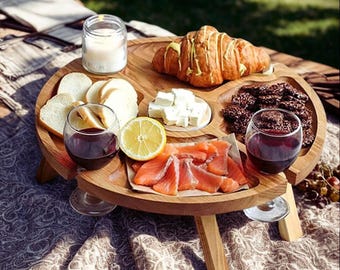 Puede incluir: Una mesa de picnic de madera con una parte superior redonda y tres patas. La mesa está llena de comida, incluyendo un cruasán, pan, queso, salmón y dos copas de vino tinto. Una vela está en un tarro de cristal.