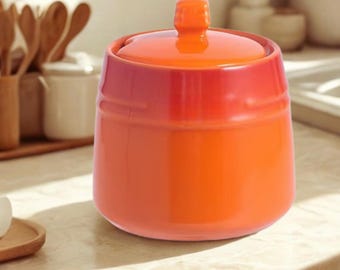 Puede incluir: Un recipiente de cerámica de color naranja brillante con tapa a juego y un pequeño pomo en la parte superior. El contenedor presenta un acabado liso y brillante, con una forma cilíndrica y una base ligeramente estrecha.