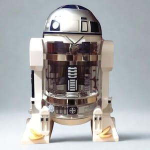 Puede incluir: Un recipiente temático de R2-D2 de Star Wars. El recipiente es blanco, plateado y transparente con detalles en negro y azul. El diseño se basa en el icónico droide, con su forma y detalles reconocibles. El recipiente tiene una sección central transparente.