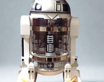Puede incluir: Un recipiente temático de R2-D2 de Star Wars. El recipiente es blanco, plateado y transparente con detalles en negro y azul. El diseño se basa en el icónico droide, con su forma y detalles reconocibles. El recipiente tiene una sección central transparente.