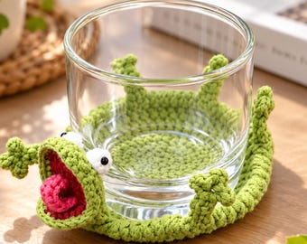 Puede incluir: Un vaso de cristal transparente descansa sobre un posavasos de ganchillo verde con forma de rana. El posavasos presenta una boca abierta con una lengua rosa, ojos blancos con pupilas negras y cuatro extremidades pequeñas.
