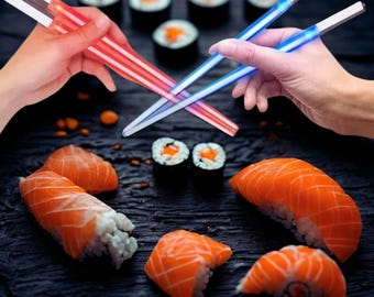 Puede incluir: Arreglo de sushi con nigiri y rollos maki, y dos pares de palillos brillantes. Un par emite luz roja, el otro azul. El sushi está dispuesto sobre una superficie oscura, creando un contraste visual.