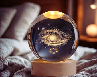 Puede incluir: Una lámpara esférica de cristal con un diseño de galaxia, colocada sobre una base de madera. La lámpara emite una luz cálida y amarilla, iluminando el patrón de la galaxia en su interior. Un cable blanco sale de la base. La lámpara está sobre una cama con una manta gris.