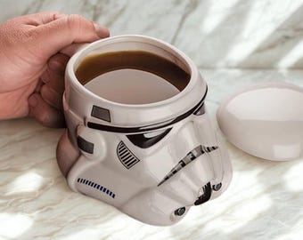 Puede incluir: Una taza de cerámica blanca con forma de casco de Stormtrooper, llena de un líquido oscuro. La taza tiene detalles negros y una tapa a juego. Una mano sostiene la taza.