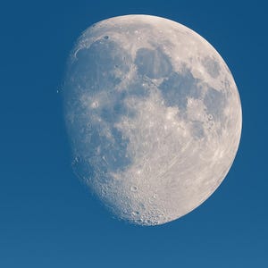 May include: A close-up of the moon against a deep blue sky. The moon is a bright, off-white color with visible craters and textured surfaces. The image captures the moon's phase, showing a partially illuminated surface.