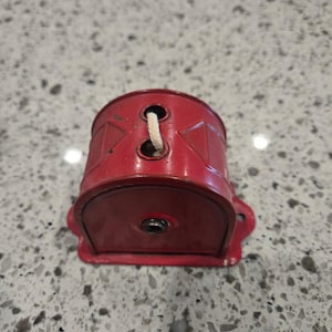Puede incluir: Un portabobinas de metal rojo vintage con un cordón blanco que pasa por dos agujeros. El portabobinas tiene una base redondeada y dos orificios de montaje. El metal muestra desgaste.