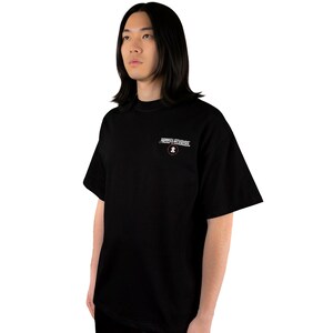 May include: Black t-shirt with the text "SONKEL STUDIOS" in white and a small circular logo. The shirt is a solid black color and has short sleeves. The model is wearing black pants.