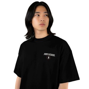 May include: Black t-shirt with the text "SONKEL STUDIOS" in white script above a small graphic. The shirt has a crew neck and short sleeves. The model is wearing the shirt.