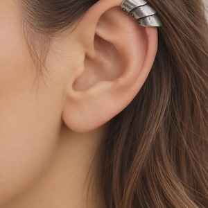 May include: A silver-toned metal ear cuff featuring a layered, segmented design that resembles armor plating. The piece has a weathered, matte finish with small circular indentations along the lower edge, securely fitted onto the upper ear cartilage.