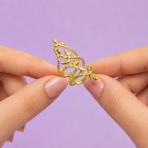 Pode incluir: Um anel de tom dourado com um design intrincado e vazado de vinhas entrelaçadas e pequenas folhas. A estrutura metálica é delicada e curva, formando uma silhueta alongada e pontiaguda que envolve o dedo com um acabamento orgânico e texturizado.
