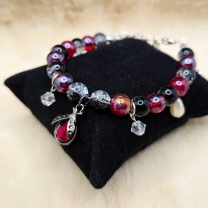 May include: A beaded bracelet featuring a mix of red, black, and iridescent beads. A silver charm in the shape of a ladybug with a red center hangs from the bracelet, along with several clear crystal charms.