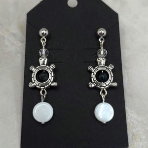 May include: Silver-tone turtle earrings. Each earring has a small silver ball, clear beads, a turtle pendant with a black centre, and a white, round bead. The earrings are displayed on a black card. The earrings are approximately 5 cm in length.